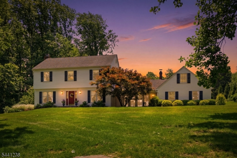 Welcome Home Embrace tranquility in this custom center-hall colonial, glowing at twilight and nestled on over two acres of lush grounds. With colorful perennials in full bloom, this home invites you to experience space, elegance, and natural beauty.